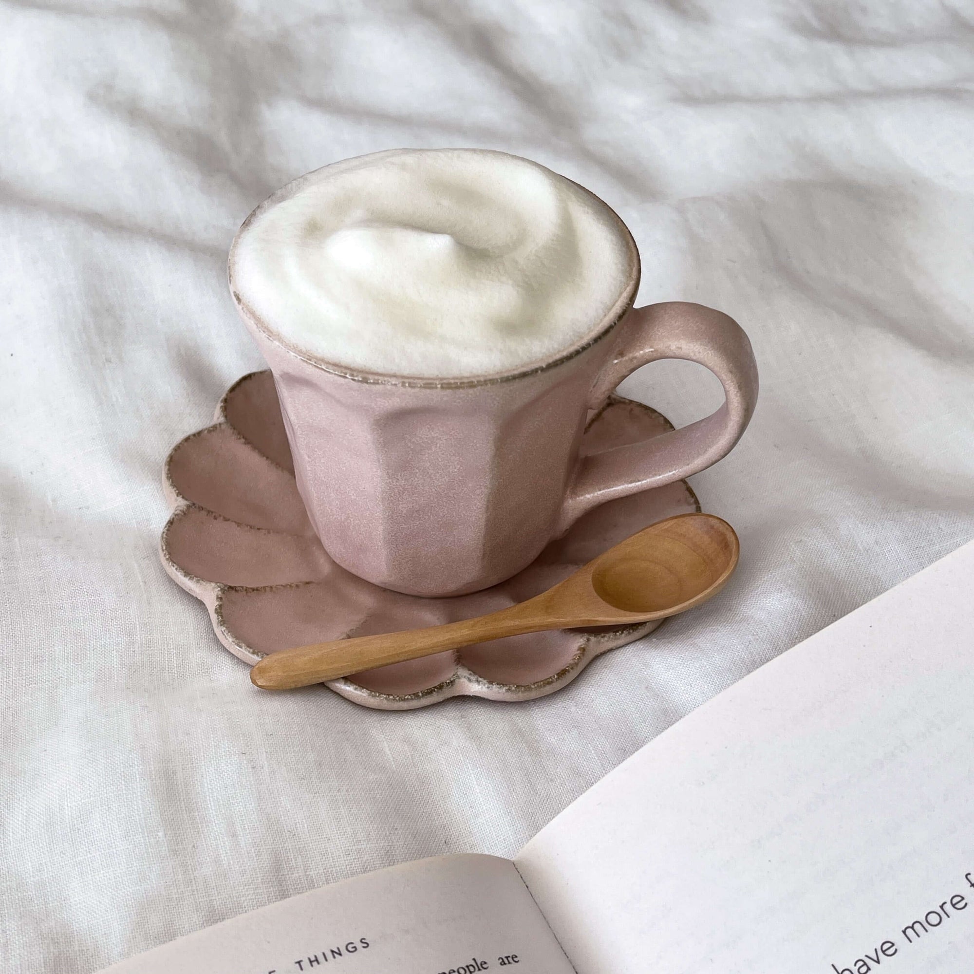 Ceramic cup with frothy milk on scalloped saucer, accompanied by a 12.1cm wooden teaspoon, placed on a white fabric background.
