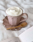 Ceramic cup with frothy milk on scalloped saucer, accompanied by a 12.1cm wooden teaspoon, placed on a white fabric background.