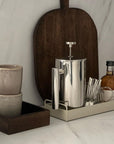Stainless steel French press coffee pot on a tray with cups and syrup on a kitchen counter setup.