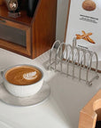 Stainless steel toast holder next to a cappuccino on a table, perfect for keeping toast warm and crisp at breakfast.