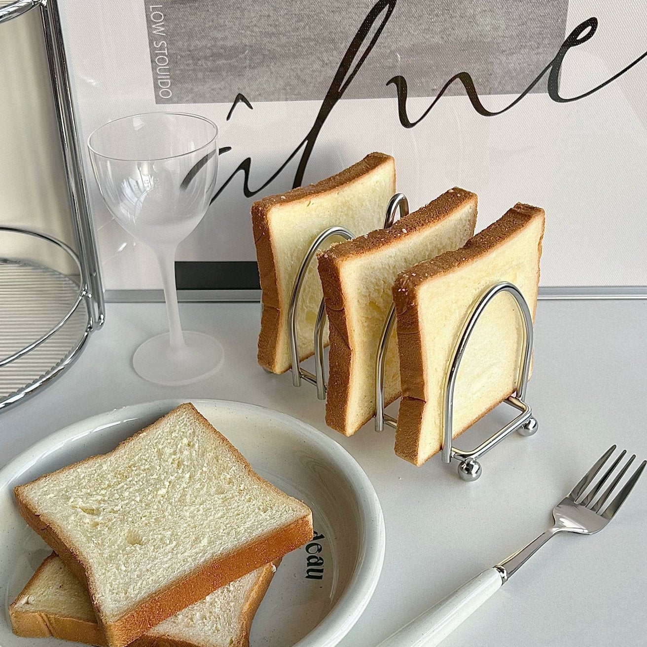 Stainless steel toast holder with six slices on a breakfast table, next to a white plate and fork, offering a stylish dining experience.