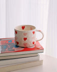 Hand-painted coffee mug with red and pink hearts, 300ml capacity, beige ceramic, placed on stacked books near a window.