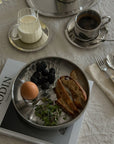 Noir Steel Plate with an egg, greens and toast on a magazine with coffee cups behind