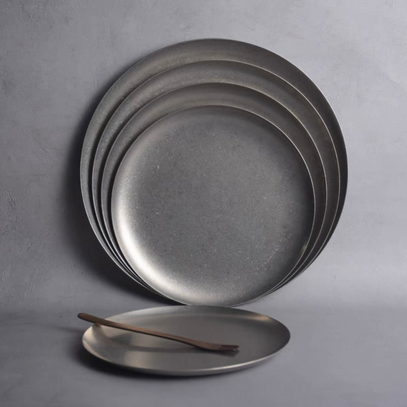 Set of matte-finish stainless steel Hakone Plates stacked with a wooden fork, perfect for elegant dining.
