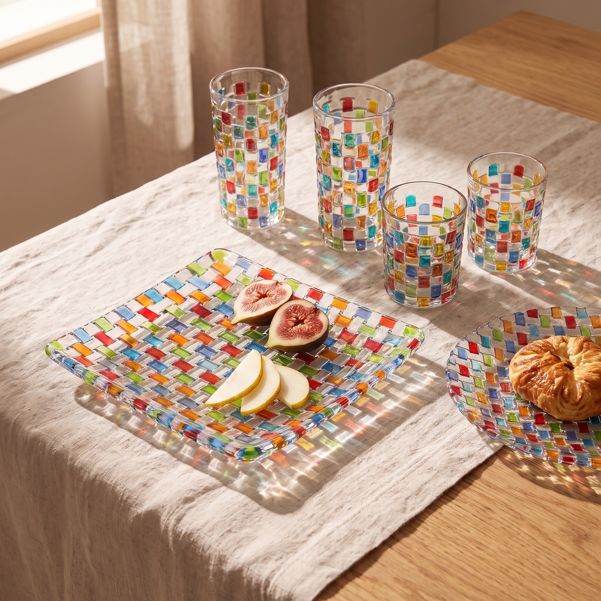 Tessera Table set, square: square platter with figs, 23.5 cm plate with a pastry and four mosaic glasses on a linen-covered table