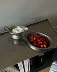 Stainless steel sauce pot with cream and bowl of strawberries on a metal surface.