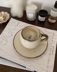 Stylish ivory mug with textured porcelain finish on a saucer, set on a wooden table beside decorative candles and a coaster.