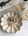 Porcelain flower-shaped jewelry dish with gold rings and pearl necklace on a marble surface alongside perfume bottles and a book.