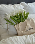 Beige tote bag with "LoveÉcru" text, filled with white tulips, resting on a plush white bedding set.
