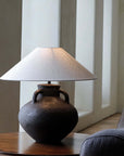 Handmade ear table lamp with pottery base and white shade, providing soft warm light on a wooden table in a cozy room.