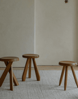 Three Meribel stools in teak wood, inspired by Alpine architecture, showcasing manual polishing and charcoal burning on a neutral rug background.