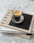 Zinc coffee glass cup with espresso on stack of Cereal magazines on marble countertop, French brasserie inspired design.