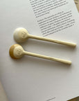 Two Marumitsu Bean Spoons with semi-matte rustic glaze tips on a book, showcasing a unique bean-shaped design and white clay finish.