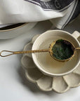 Brass handwoven tea strainer with loose leaf tea over a ceramic cup on a scalloped saucer.