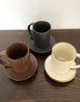 Three textured porcelain mugs on saucers, featuring black, brown, and cream colors, placed on a wooden surface.