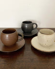 Three textured porcelain mugs and saucers in brown, black, and cream, displayed on a wooden surface.