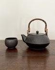 Black textured teapot with wicker handle next to a small black cup on a wooden table.